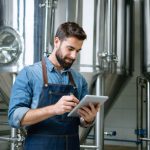 Craft brewer in a denim apron holding a tablet with a blank glowing screen inside a modern stainless-steel brewhouse, eye-level three-quarter view with soft natural light and blurred tanks, hoses, and kegs in the background.