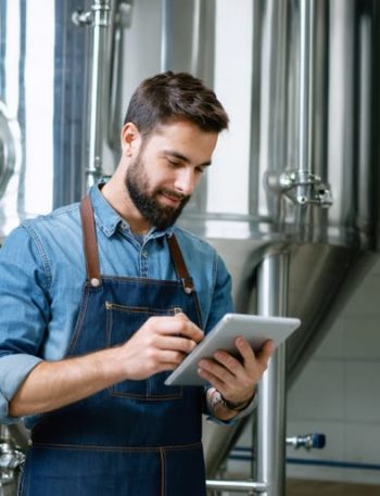 Craft brewer in a denim apron holding a tablet with a blank glowing screen inside a modern stainless-steel brewhouse, eye-level three-quarter view with soft natural light and blurred tanks, hoses, and kegs in the background.