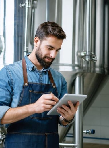 Craft brewer in a denim apron holding a tablet with a blank glowing screen inside a modern stainless-steel brewhouse, eye-level three-quarter view with soft natural light and blurred tanks, hoses, and kegs in the background.