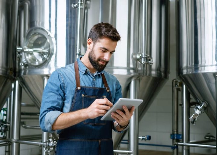 Craft brewer in a denim apron holding a tablet with a blank glowing screen inside a modern stainless-steel brewhouse, eye-level three-quarter view with soft natural light and blurred tanks, hoses, and kegs in the background.