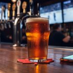 Close-up of a pint of amber craft beer on a red maple-leaf coaster beside a smartphone on a bar, with blurred hockey game on TV screens and tap handles in the background; no visible text or logos.