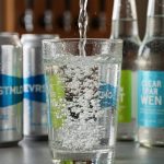 Close-up of condensation-covered sparkling water in a glass with visible bubbles, with chilled cans in the foreground and a blurred craft taproom background.