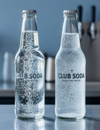 Two clear bottles of carbonated beverages—club soda and sparkling water—on a bar counter with bubbles rising.