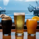 Coastal bar counter at dusk with three local drinks—an oyster stout, salt-rimmed lager, and tropical IPA—framed by a window view of the ocean.