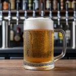 Frosty beer mug with condensation and a golden lager pour on a wooden bar, with out-of-focus beer taps and bottles in the background.